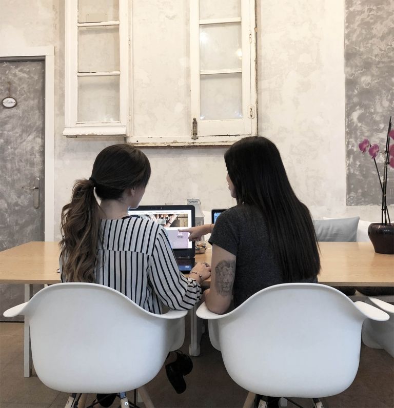 fotografía de dos chicas de espaldas sentadas a una mesa trabajando en sus portátiles, parece que estén compartiendo ideas