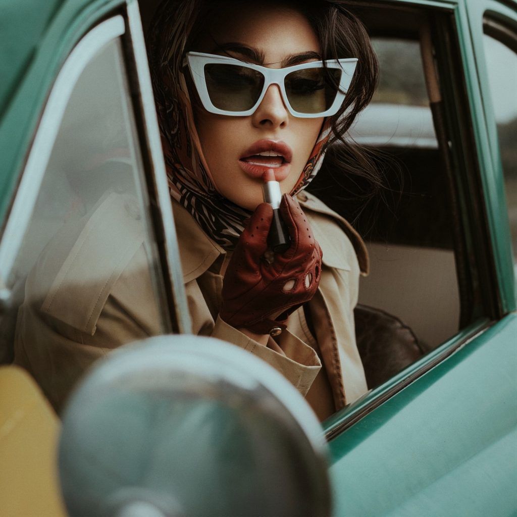 Mujer bonita con aire vintage pintándose los labios utilizando de espejo el retrovisor del coche donde está sentada