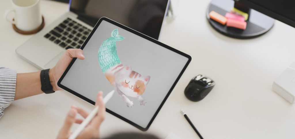 Vemos una mesa de trabajo donde las manos de una persona sujetan un apple pencil y un ipad donde está realizando una ilustración infantil de un gato con cuerpo de sirena