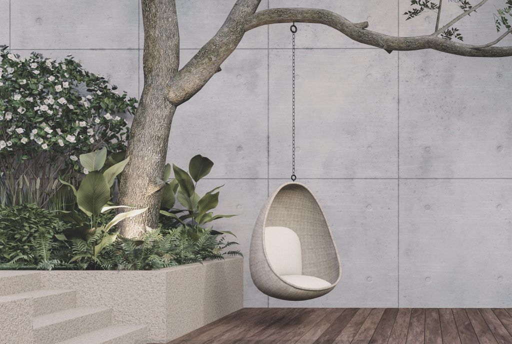 Espacio exterior, vemos una pared de hormigón, suelo de madera, a la izquierda una jardinera con plantas y un árbol grande que sujeta un asiento de columpio con forma de huevo con cojines blancos