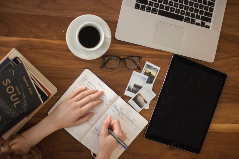 Vemos una fotografía tomada desde arriba donde vemos las manos de una persona tomando apuntes en un cuaderno, rodeada de libretas, libros, fotografías, una taza de café y un portátil