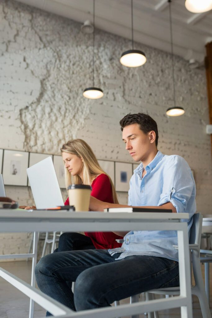 En la imagen vemos a una chica y un chico sentados a una mesa trabajando en sus respectivos portátiles, el espacio es industrial y un tanto frío