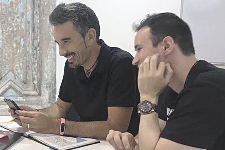 Dos profesores de Artes Creativas sentados a una mesa con cuadernos, muy sonrientes y vestidos con camisetas negras
