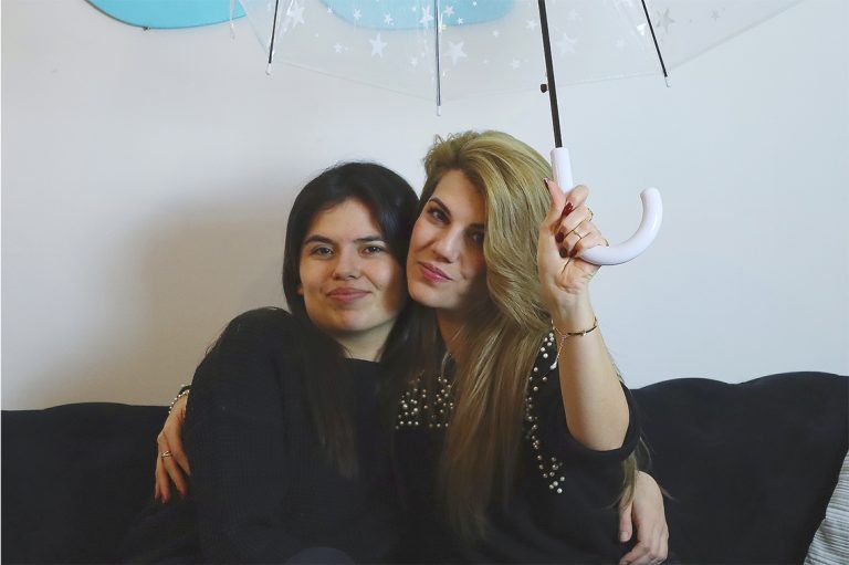 Dos profesoras de Artes Creativas sentadas en un sofá negro, parecen amables, se abrazan y una de ella sujeta un paraguas abierto sobre ellas