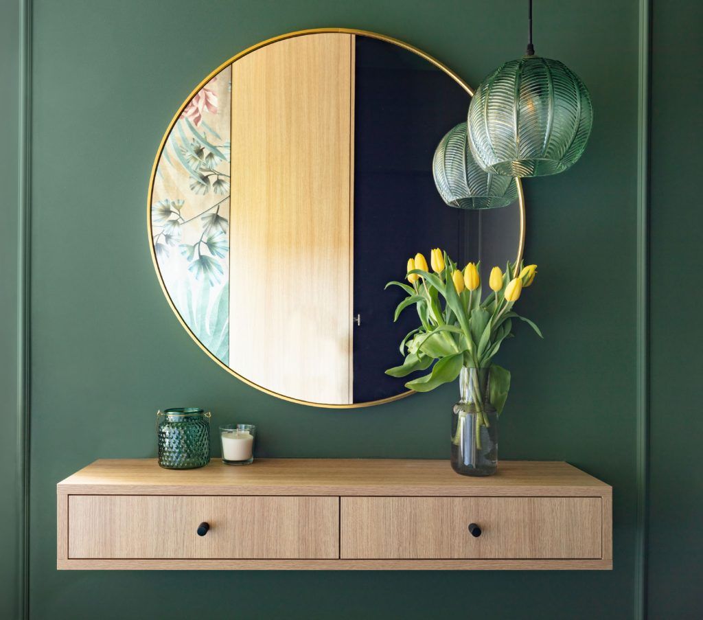 Fotografía frontal de pared verde oscura donde vemos un mueble de madera, un espejo redondo, adornos de cristal verdoso, velas, un jarrón con tulipanes amarillos y una lámpara de cristal verde muy bonita. El espacio es moderno y agradable