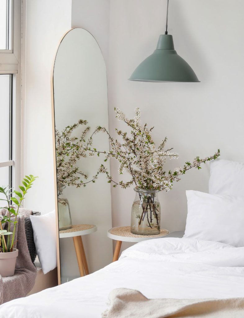 Espacio de dormitorio muy agradable, sábanas blancas, junto a la cama vemos una lámpara industrial de techo color verde menta, debajo una mesita que alberga un gran jarrón con flores silvestres.