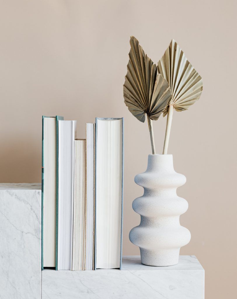 Fotografía de un detalle decorativo de unos libros junto un jarrón con unas horas doradas sobre un soporte de mármol.