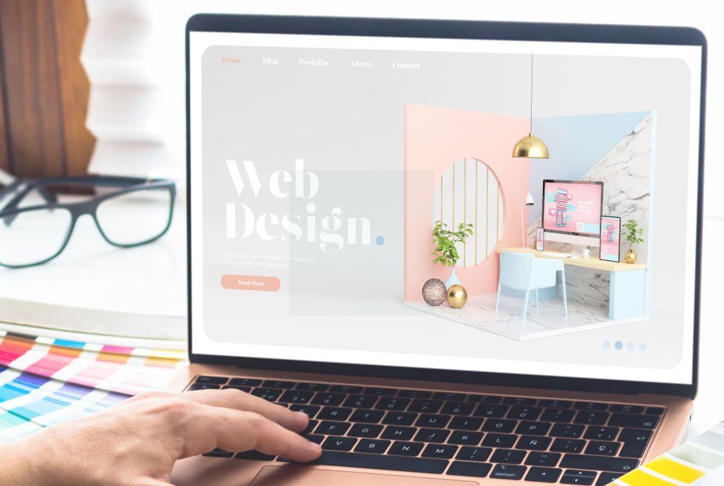 Vemos la mano de una persona sobre el teclado de un portátil donde podemos ver en la pantalla una fotografía de un espacio de interior de oficina con tonalidad muy agradable, es un espacio chic y acogedor, con unas letras en grande donde se lee Web Design. Detrás del portátil podemos observar unas gafas de pasta y unas pantoneras de color por encima del escritorio.