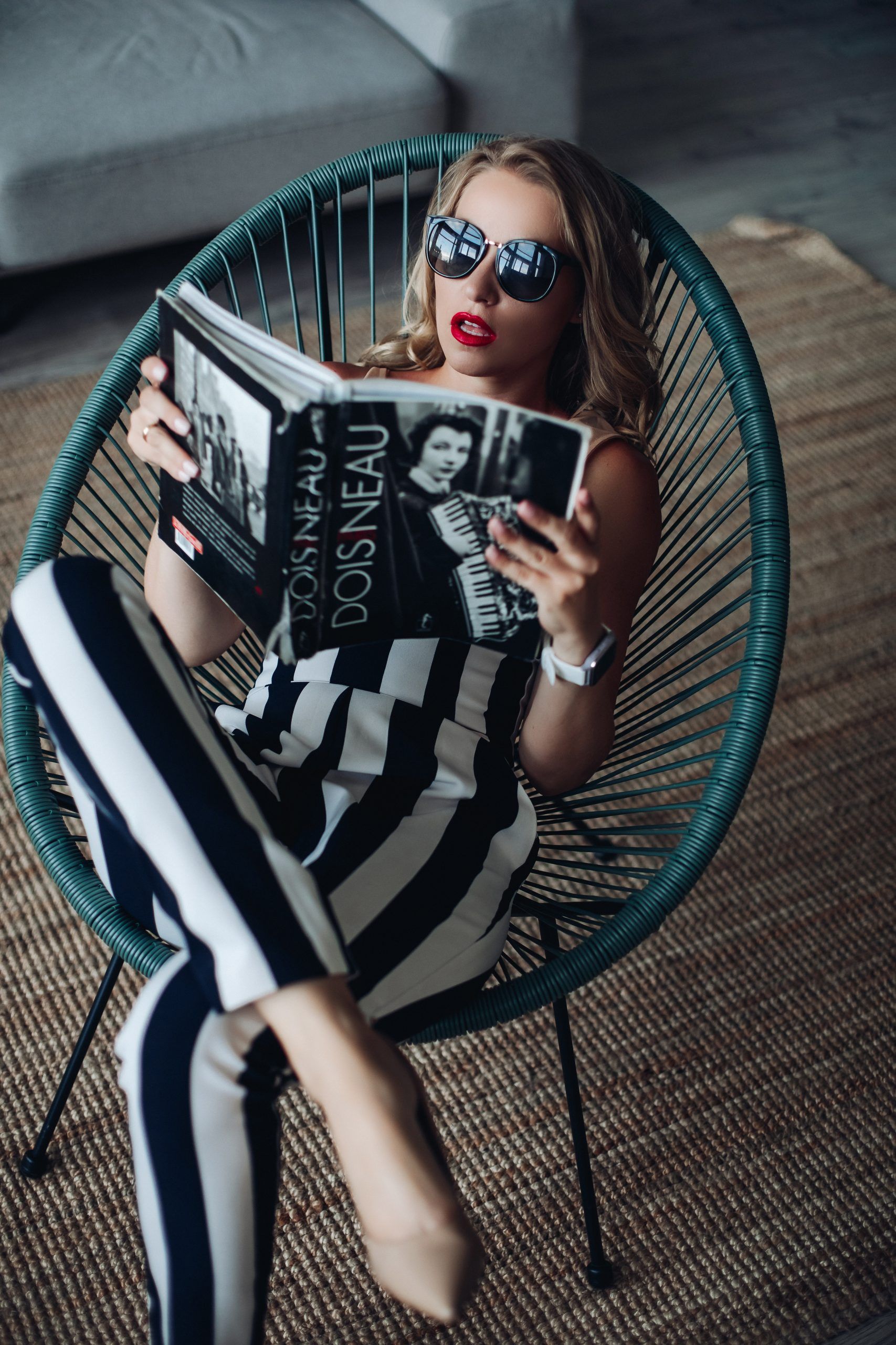 Chica atractiva sentada en una silla de diseño, lleva gafas de sol grandes, labios pintados de rojo, pelo suelto rubio, viste tacón y un mono de rayas blancas y negras anchas, sujeta una revista vintage en color blanco y negro